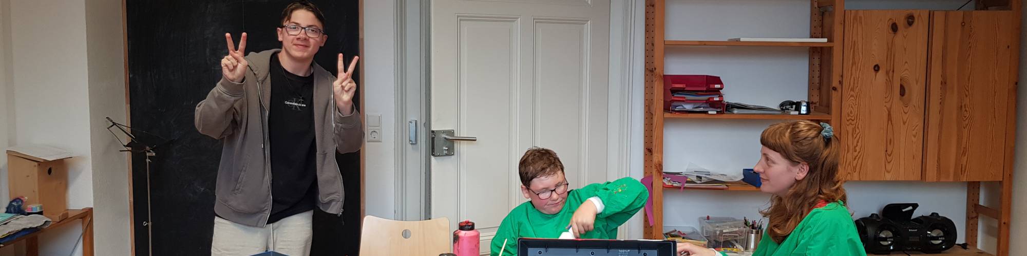 Ein Jugendlicher zeigt ein Friedenszeichen, während zwei jüngere Personen an einem Tisch sitzen und an einem Projekt arbeiten. Ein Raum mit Regalen im Hintergrund