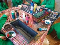 Zwei Kinder, die grüne Malkittel tragen, sind dabei, ein Objekt rot und weiß zu bemalen. Um sie herum sind verschiedene Farben, Pinsel und Becher mit Flüssigkeit zu sehen.